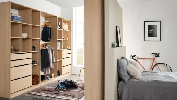 Un armario moderno y funcional, ideal para aprender como organizar un armario. Ropa colgada, cajones y estantes ordenados, con una bicicleta naranja y una cama al fondo.