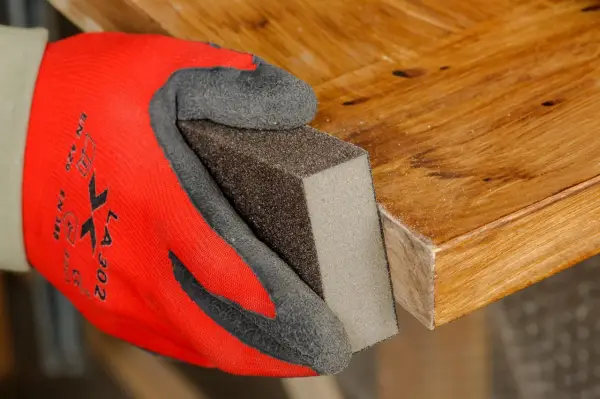 Mano con guante rojo lijando borde de puerta de madera para rebajarla.