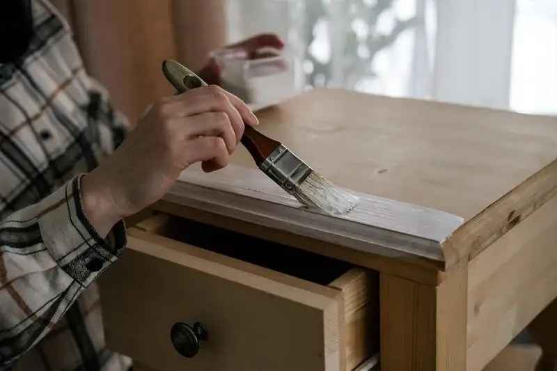 Mano con brocha pintando de blanco la madera de un mueble.